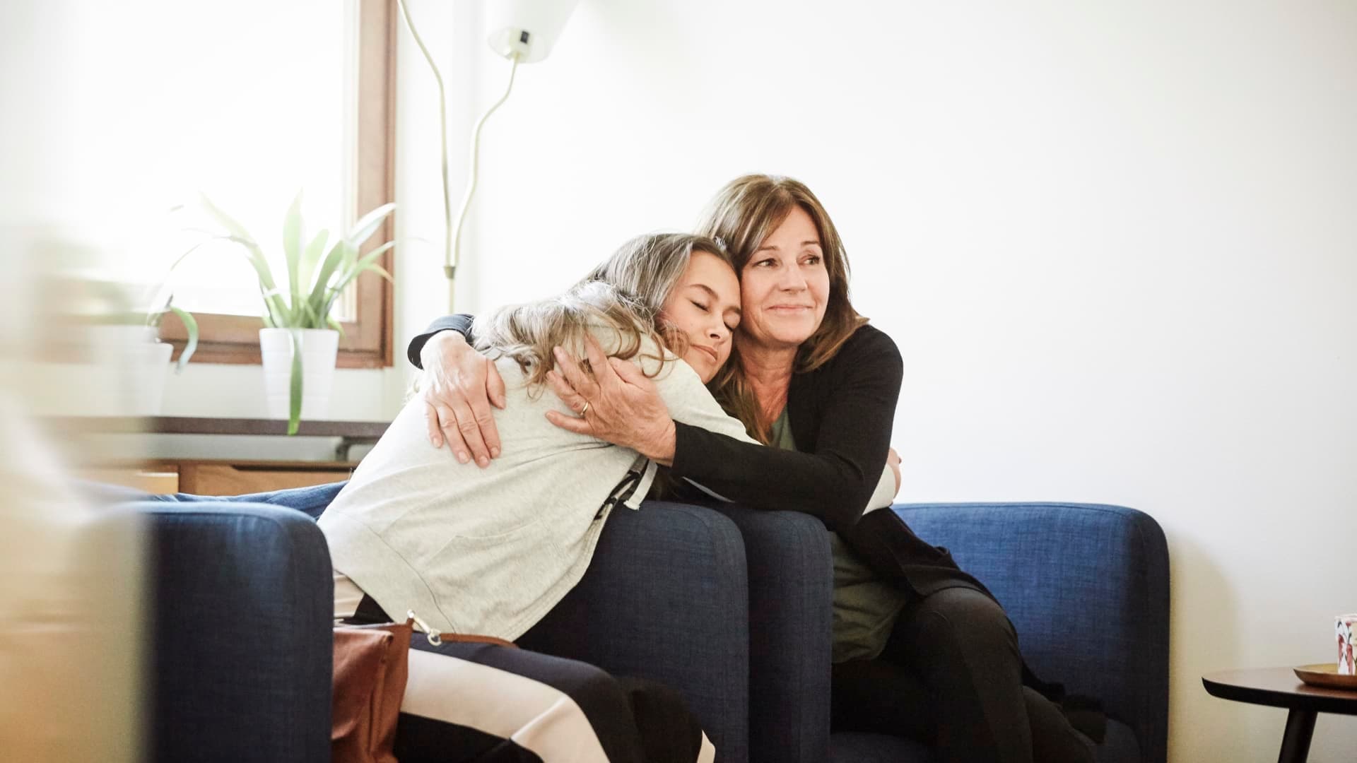 A woman gives a comforting hug to a young girl sitting on a blue sofa.