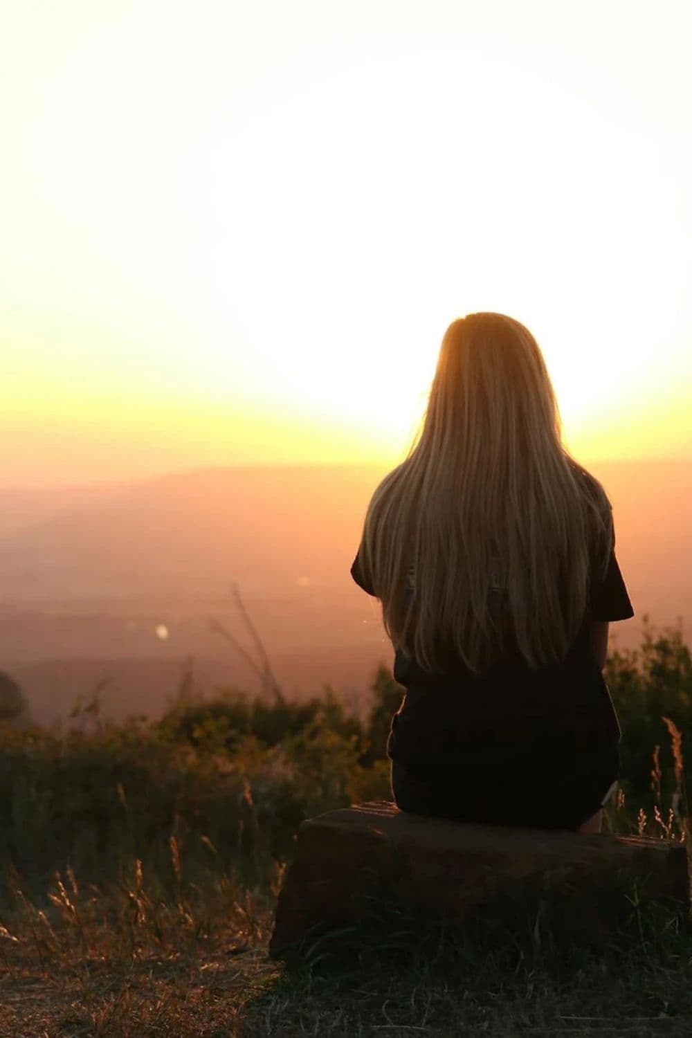 Woman with long blonde hair sits on a rock, watching a golden valley sunset.