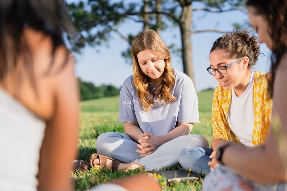 Young women sitting in a circle on a grassy field, praying together with heads bowed.