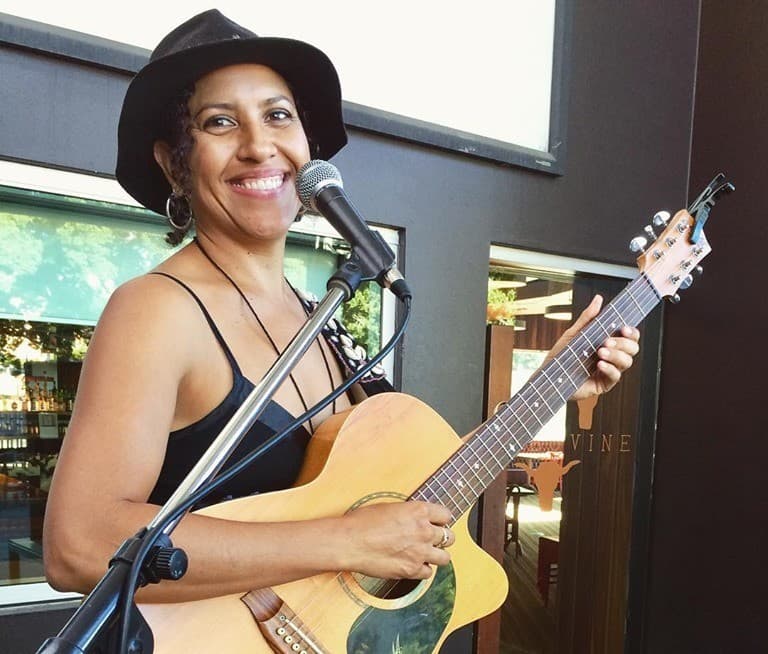 Smiling female musician in a black hat playing an acoustic guitar at a microphone.