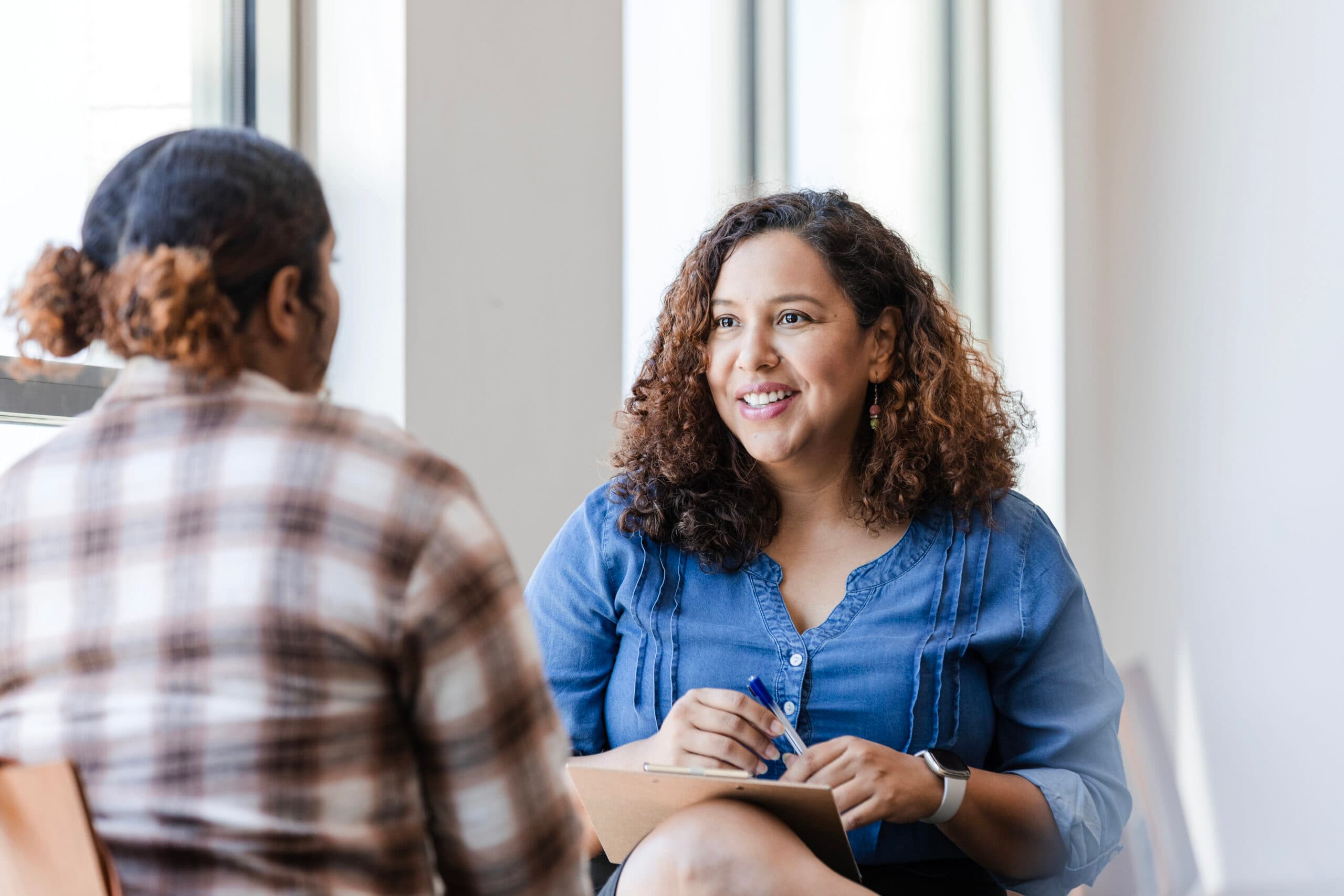 Smiling woman with curly hair holding a clipboard and pen while talking to another woman.