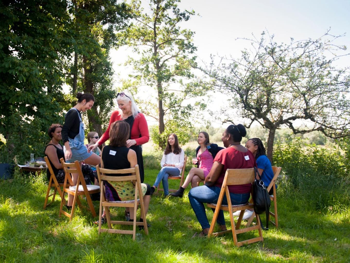 Diverse women sit in a circle on wooden chairs in a sunny, grassy field.