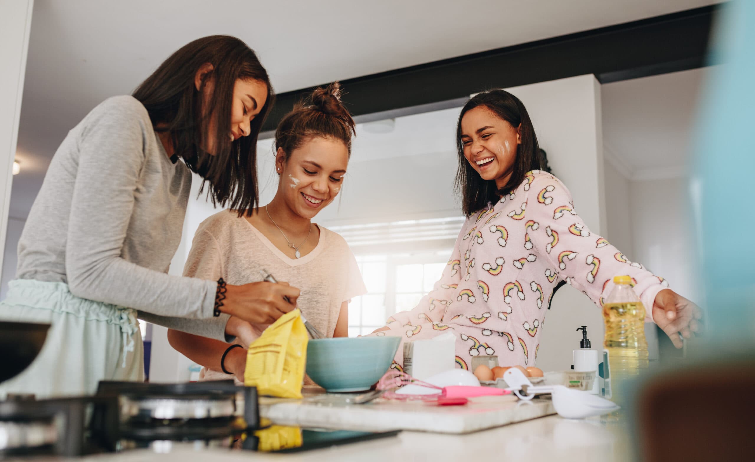 Three friends laugh while baking together in a kitchen with flour on their faces.