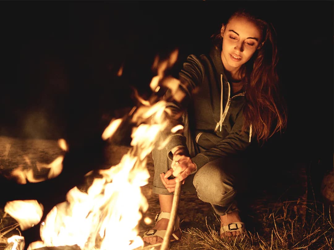 Young woman in a hoodie crouches by a bright campfire at night, holding a stick.