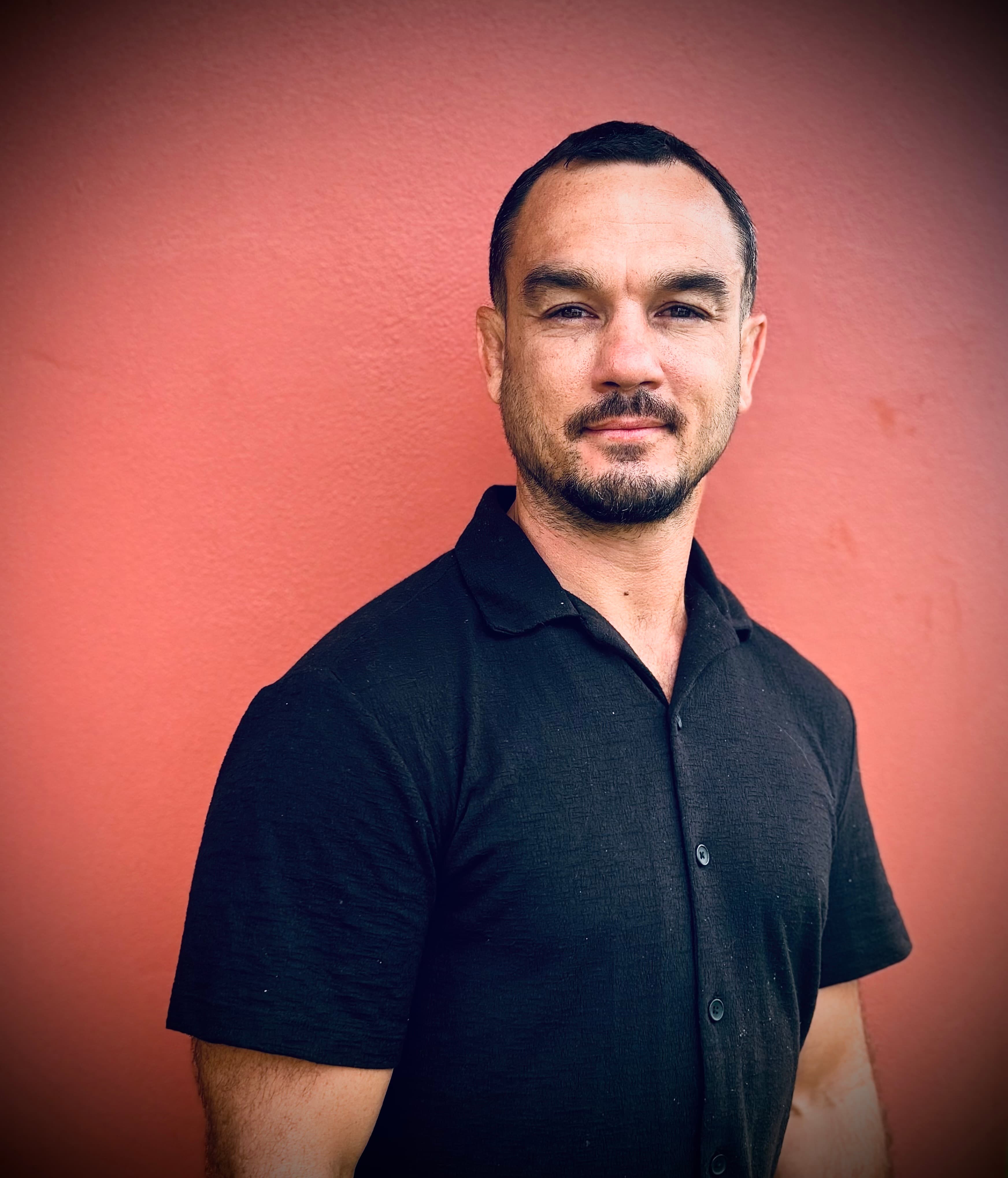 Man with a goatee and mustache in a black shirt against a textured reddish-pink wall.