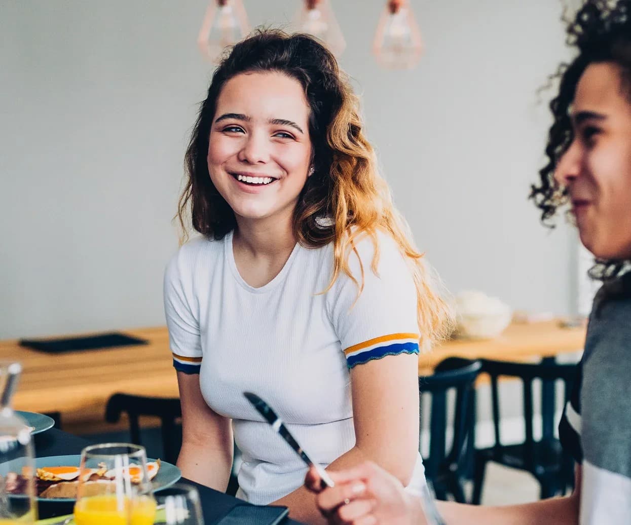 A happy young woman with wavy hair smiles while sitting at a breakfast table with friends.