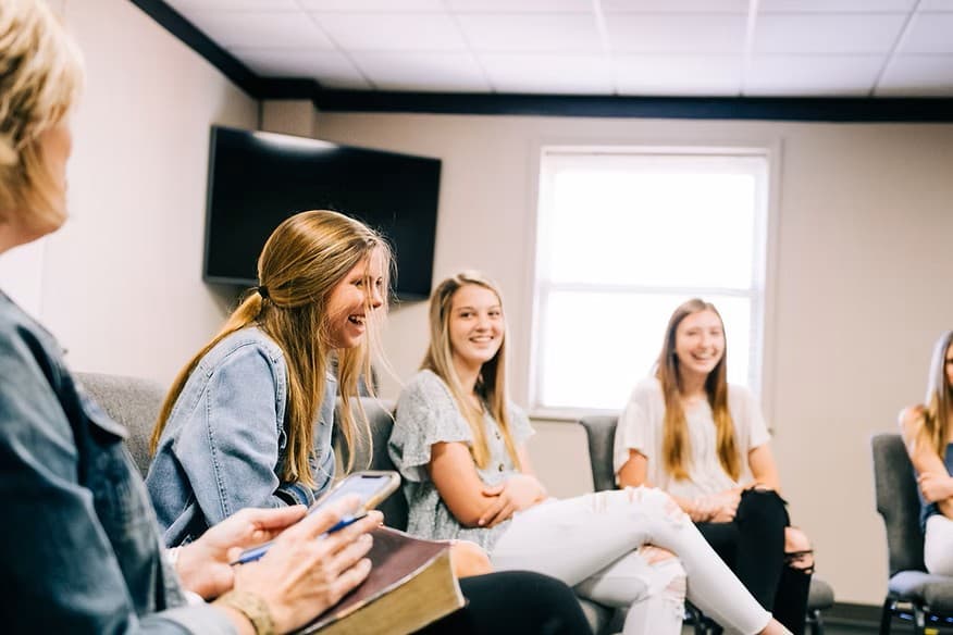 Young women sitting in a circle, smiling and laughing during a group discussion indoors.