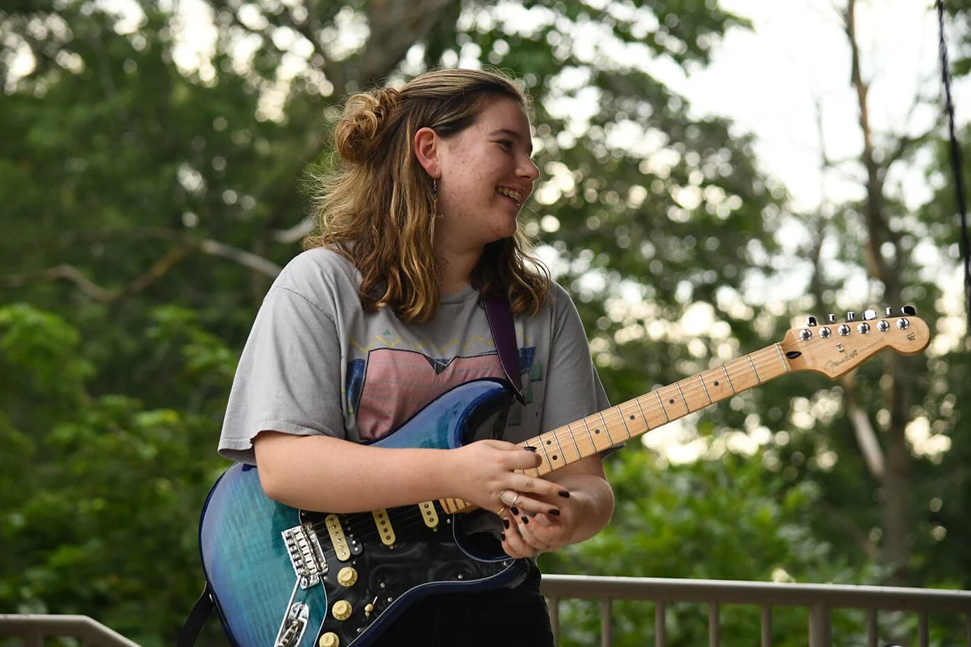 Smiling young woman with light brown hair holds a blue electric guitar against green trees.