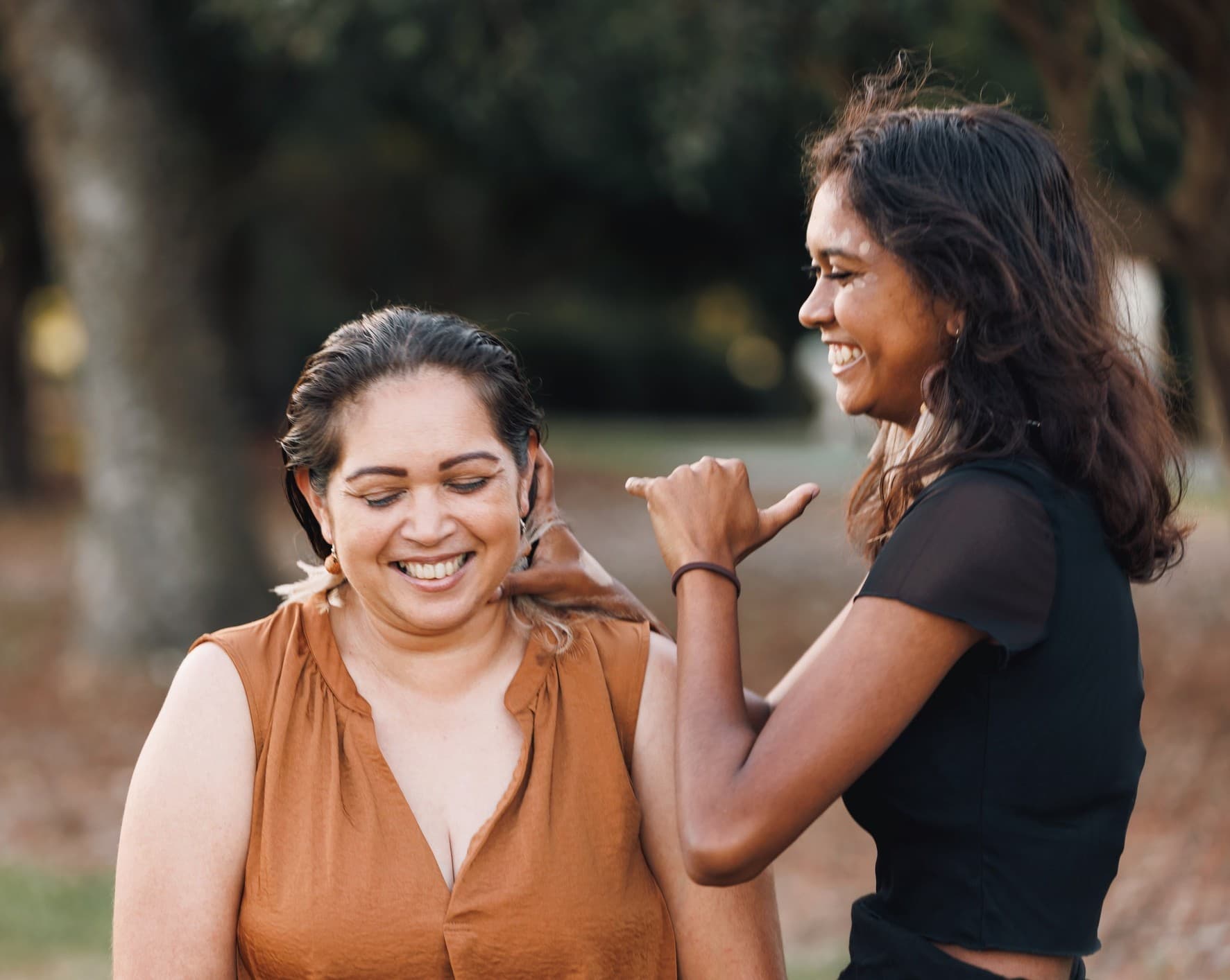 Two women smiling outdoors, one with vitiligo affectionately touching the other's hair and neck.