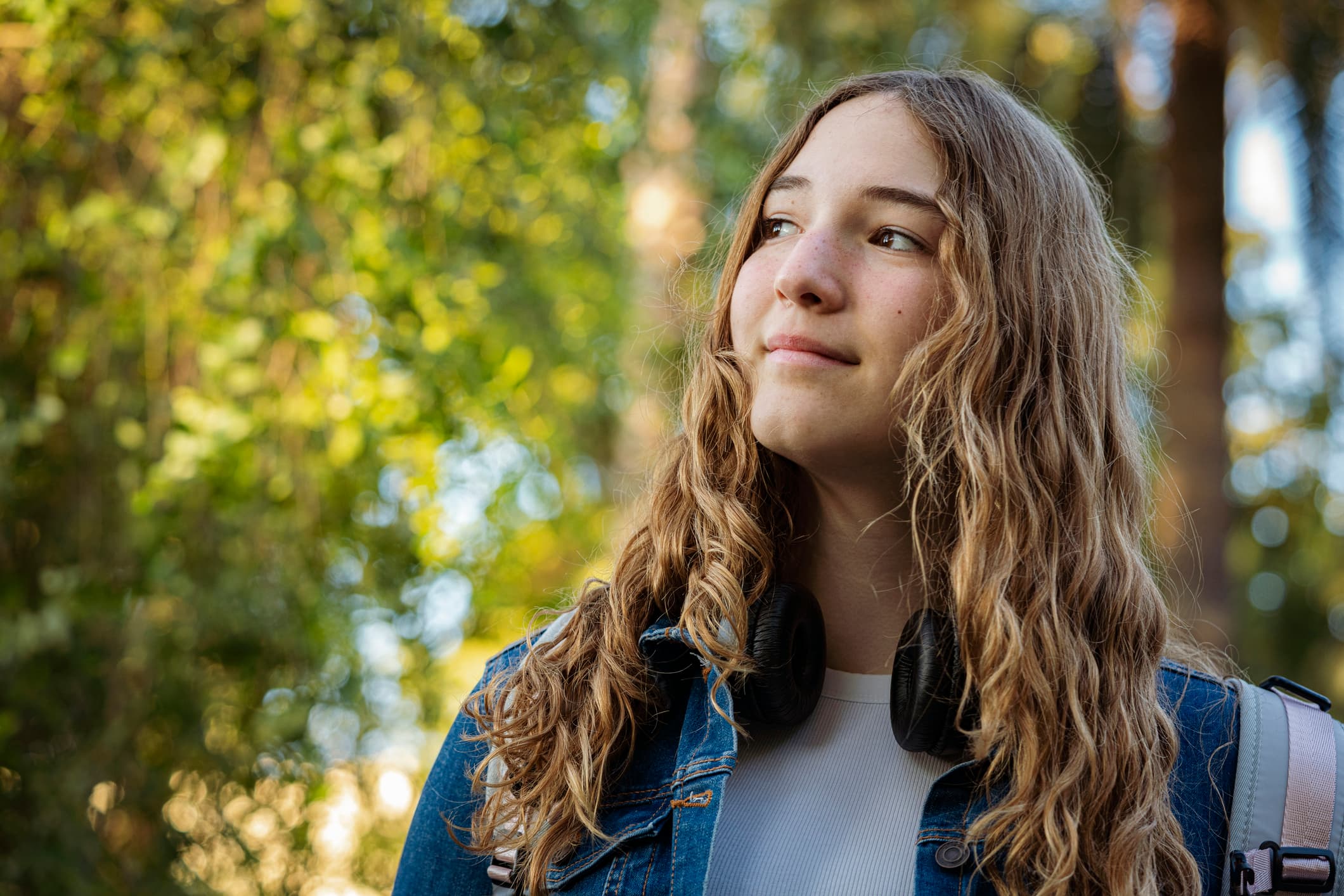 Smiling young woman with wavy hair and headphones around her neck in a sunlit park.