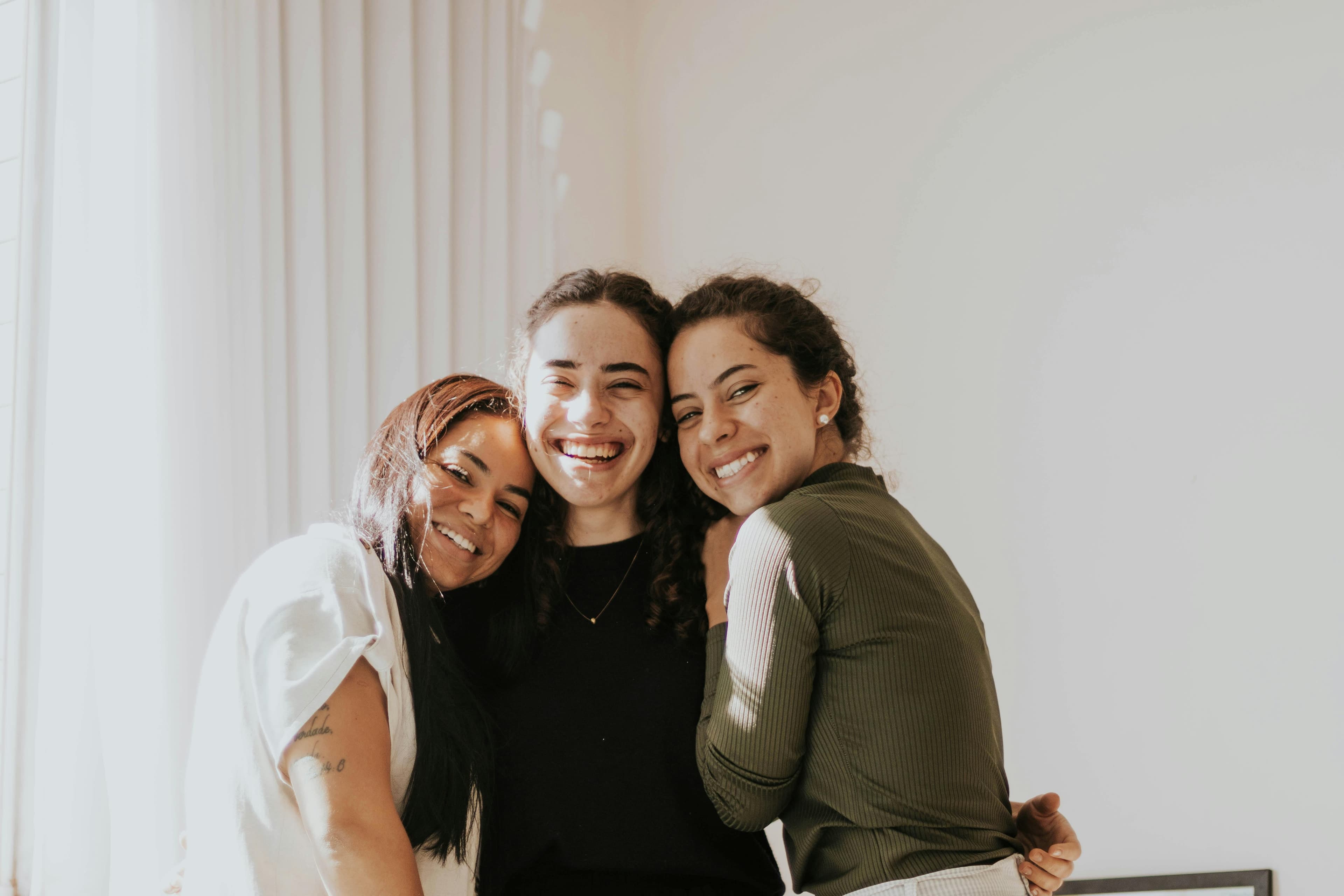 Three young women with joyful expressions hugging each other in a brightly lit room.