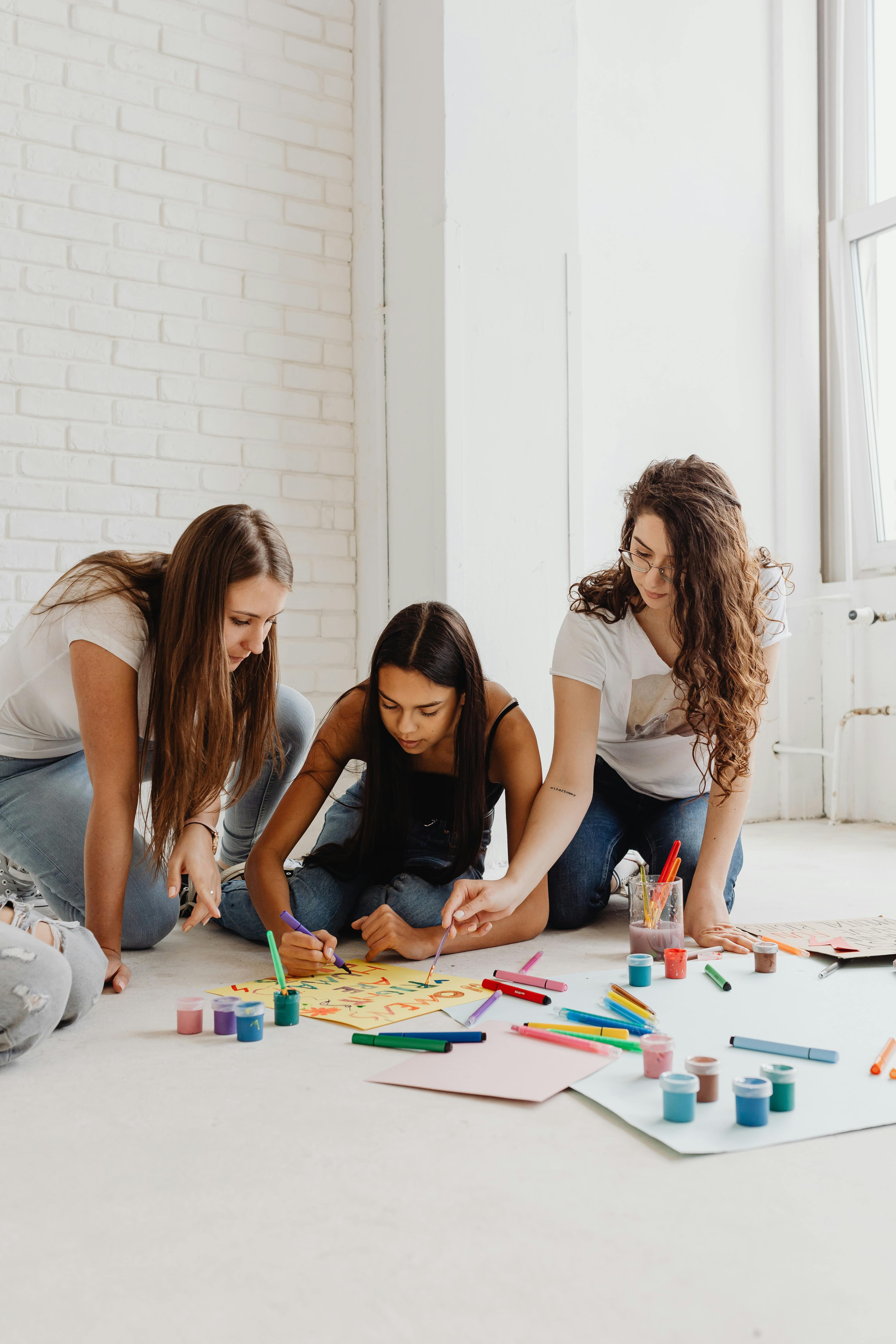 Three young women sit on the floor, painting and drawing together with colorful art supplies.