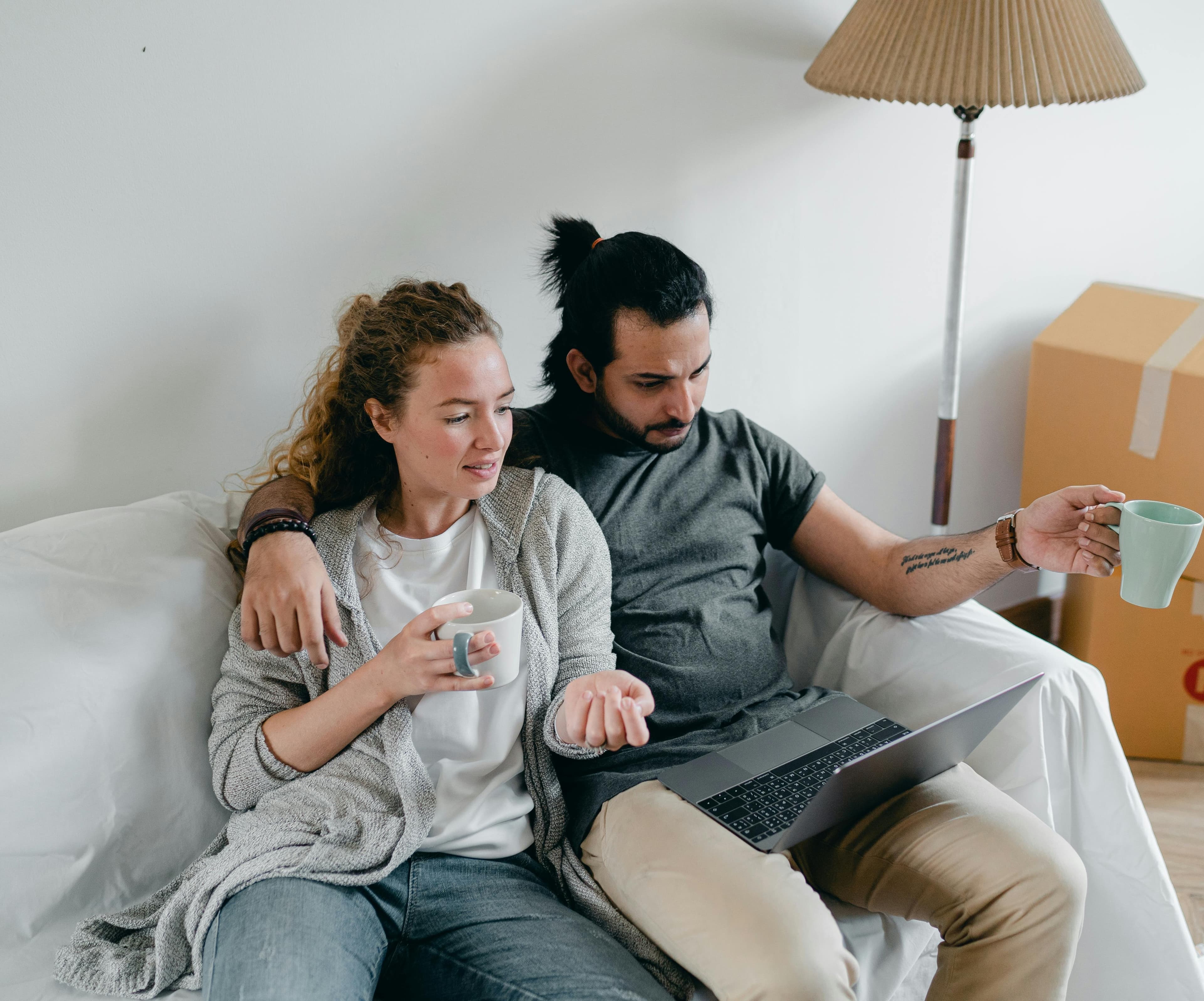 Couple on a couch with coffee mugs, looking at a laptop amidst cardboard moving boxes.