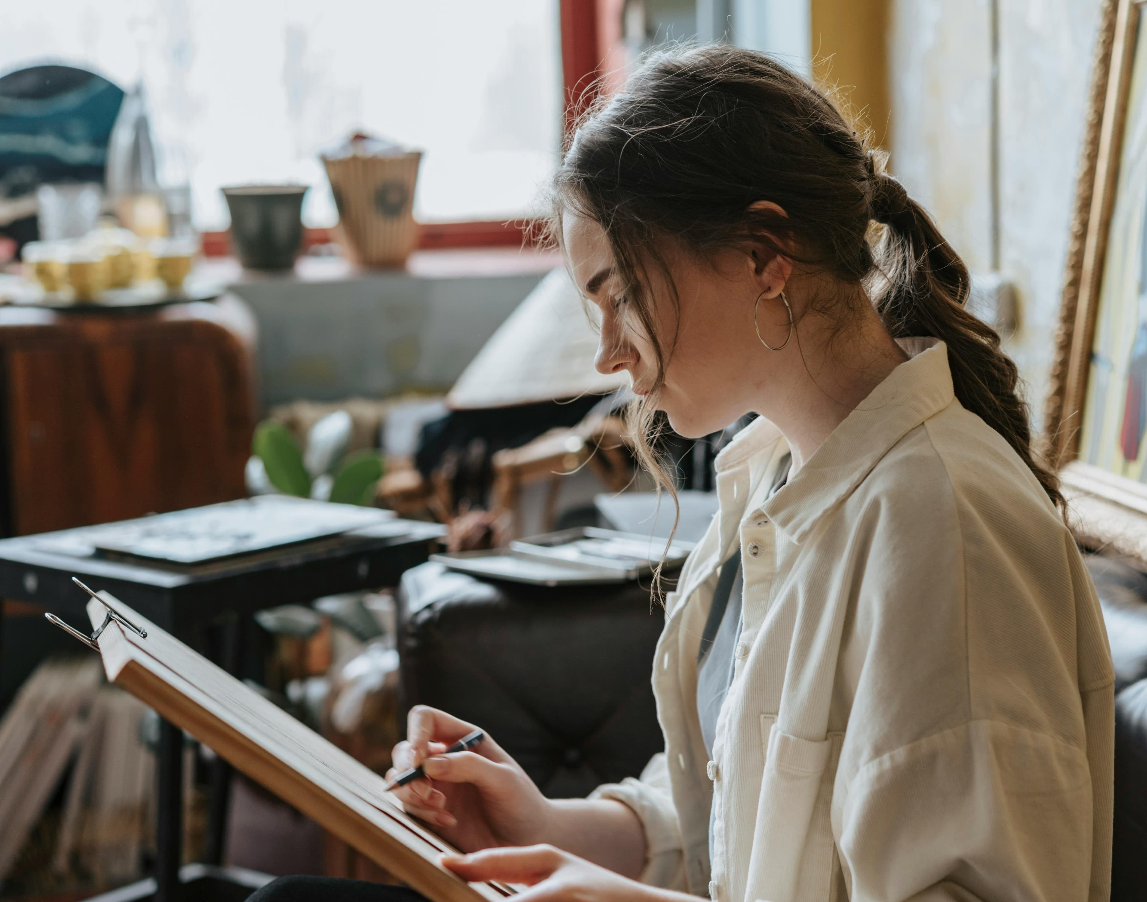 Young woman with a ponytail sketching on a wooden drawing board in a cluttered studio.