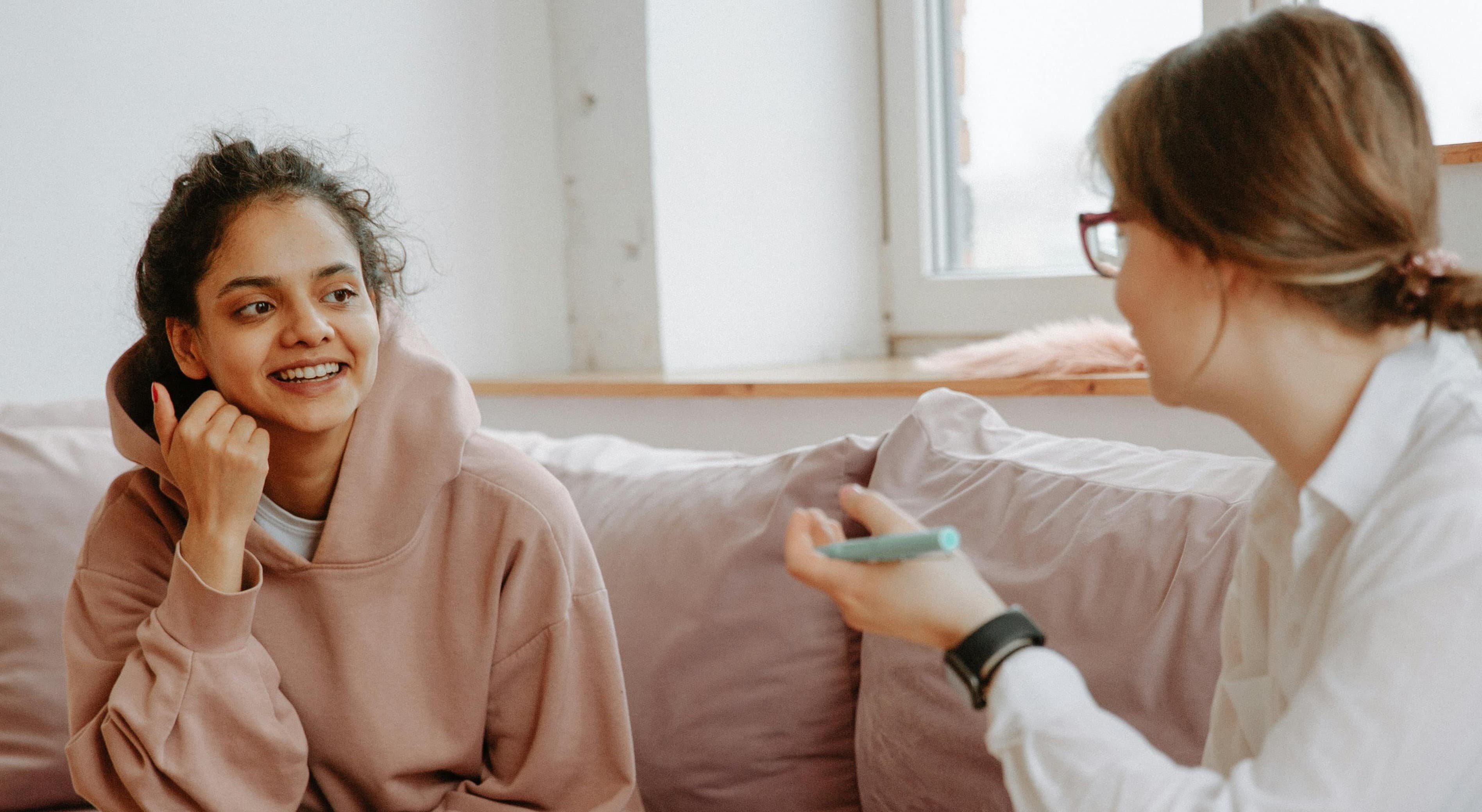 Smiling woman in a pink hoodie listens intently to another woman gesturing with a pen.