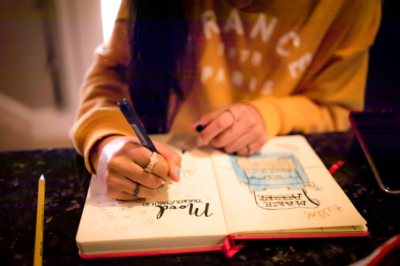 A person in a yellow sweatshirt draws a mood tracker in an open bullet journal.